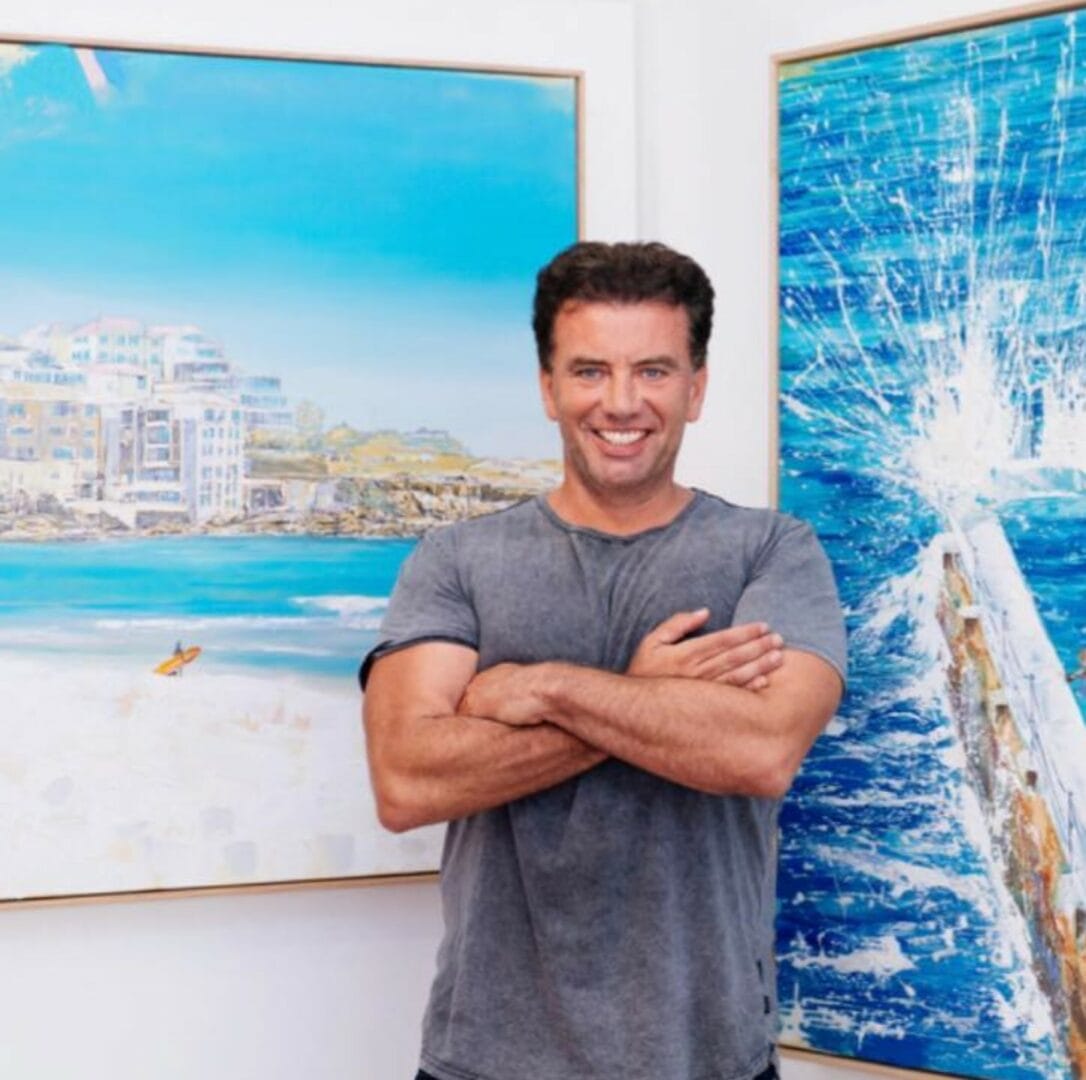 Man standing with arms crossed in front of two coastal-themed paintings